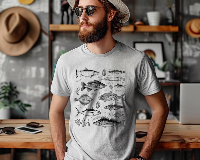 Puede incluir: Camiseta gris claro con una ilustraci&oacute;n detallada en blanco y negro de varias especies de peces. El dise&ntilde;o incluye diferentes tipos de peces, algas marinas y peque&ntilde;as plantas acu&aacute;ticas. Una prenda casual para el d&iacute;a a d&iacute;a.