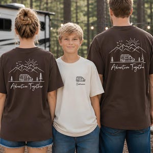 May include: Three people wearing matching t-shirts with a camping theme. The shirts are brown and cream-colored, featuring a mountain, sun, and camper van graphic with the words "Adventure Together" and "Hometown". The shirts are perfect for family camping trips.