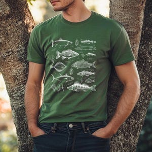 Puede incluir: Camiseta verde con una ilustraci&oacute;n blanca detallada de varias especies de peces. El dise&ntilde;o incluye varios peces y plantas acu&aacute;ticas. La camiseta es de color verde liso y se lleva con vaqueros azules.