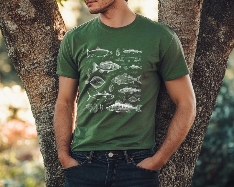 Puede incluir: Camiseta verde con una ilustraci&oacute;n blanca detallada de varias especies de peces. El dise&ntilde;o incluye varios peces y plantas acu&aacute;ticas. La camiseta es de color verde liso y se lleva con vaqueros azules.