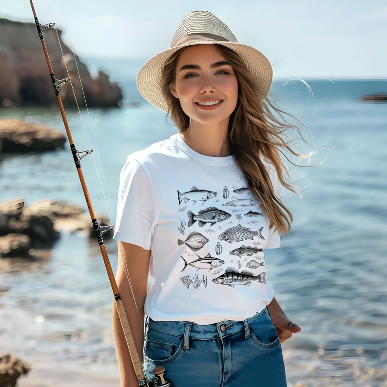Puede incluir: Una camiseta blanca con una ilustraci&oacute;n en blanco y negro de varias especies de peces. La persona lleva un sombrero de paja y vaqueros azules, sosteniendo una ca&ntilde;a de pescar cerca del mar.