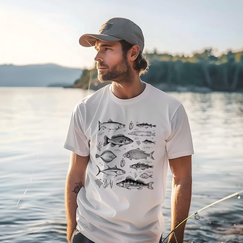 Puede incluir: Camiseta blanca con una ilustraci&oacute;n en blanco y negro de varias especies de peces. El dise&ntilde;o incluye diferentes tipos de peces, con elementos bot&aacute;nicos. La camiseta la lleva una persona con una gorra gris.