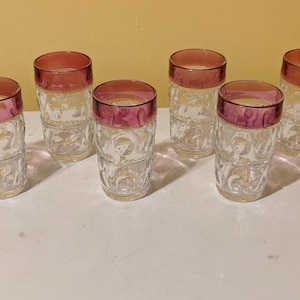 Puede incluir: Un juego de seis vasos de vidrio transparente, cada uno con un patrón texturizado en relieve en la parte inferior y un borde liso de color rosa intenso. Los vasos tienen forma cilíndrica con una base plana, diseñados para servir bebidas.