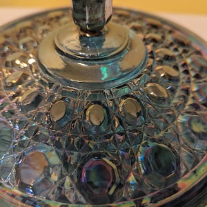May include: A round, iridescent blue glass lid featuring a central knob handle and a textured surface with a repeating pattern of raised, faceted circular shapes and geometric cross-hatching, reflecting light with metallic rainbow hues.