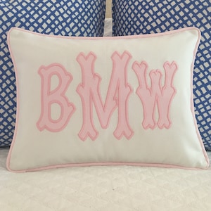 May include: A rectangular white pillow with pink trim and the embroidered letters "BMW" in a decorative font. The pillow is on a white quilted surface, with blue patterned pillows in the background.