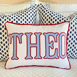 Pode incluir: Uma almofada retangular branca com o nome THEO em letras grandes azul-claras com contorno bordado a vermelho. A almofada tem um debrum vermelho e uma textura subtil em losangos, posicionada &agrave; frente de duas almofadas com padr&atilde;o azul e branco.