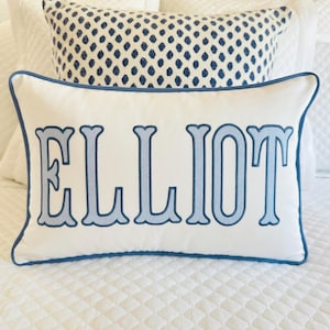 Pode incluir: Uma almofada retangular branca com o nome ELLIOT bordado em azul claro e um contorno azul escuro. A almofada tem um acabamento em tecido texturizado e uma borda com debrum azul escuro, posicionada à frente de uma almofada com padrão azul e branco.