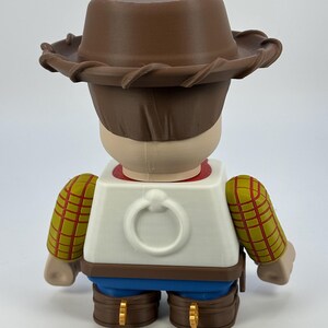 May include: A figurine of a character from the animated film Toy Story, viewed from the back. The figure wears a brown cowboy hat, a white vest with a ring detail, and blue pants. The sleeves are yellow with a red plaid pattern.