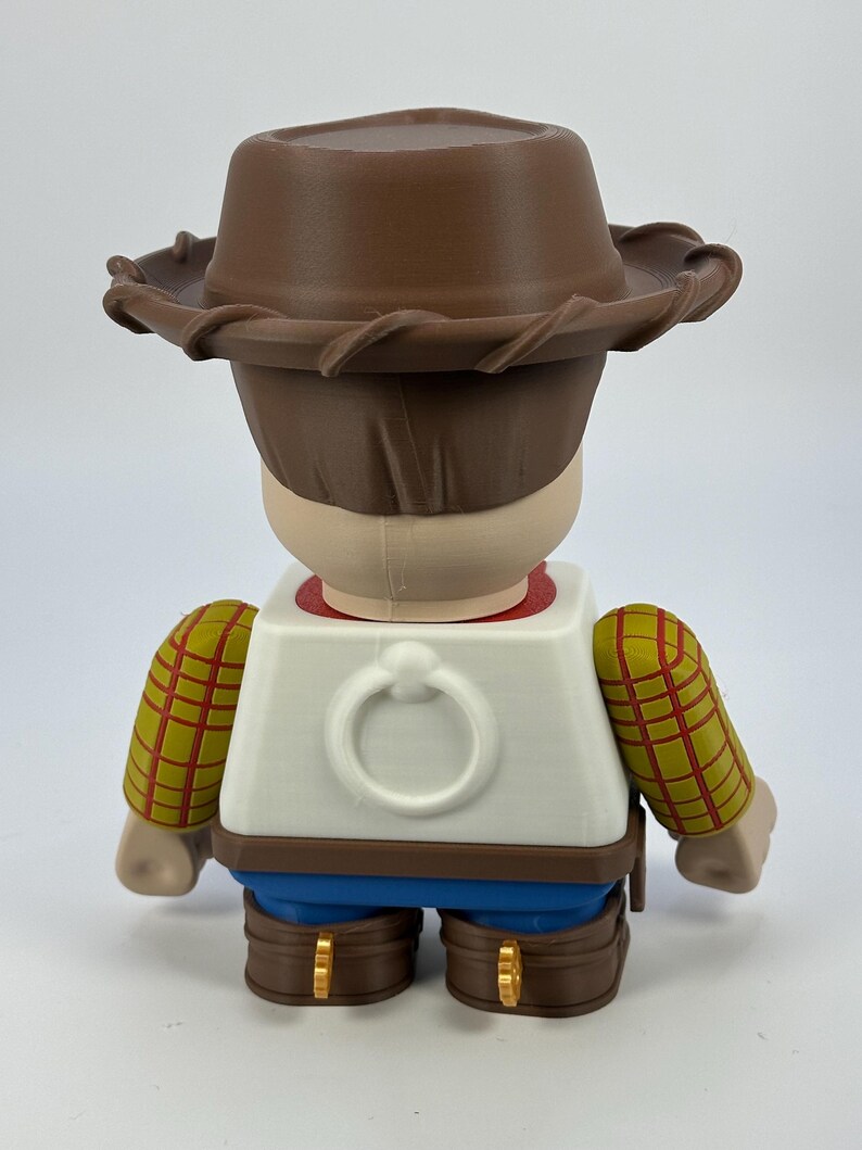 May include: A figurine of a character from the animated film Toy Story, viewed from the back. The figure wears a brown cowboy hat, a white vest with a ring detail, and blue pants. The sleeves are yellow with a red plaid pattern.