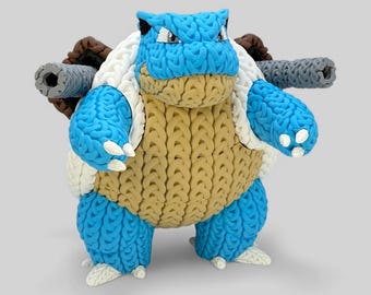 May include: A handmade, blue, white, and tan Blastoise figure. The figure has two gray cannons on its back, white claws, and a tan chest. The figure is made of a woven material and stands on a light gray surface.