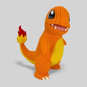 May include: A knitted orange Charmander figure with yellow accents on its belly and tail. The figure has white teeth, a pink mouth, and a red and yellow flame on its tail. The eyes are black and white.