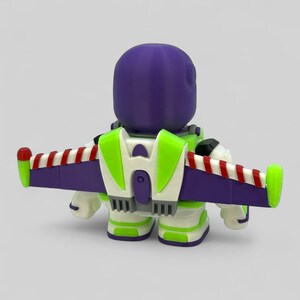 May include: A toy figure of Buzz Lightyear, seen from behind. The figure has a purple helmet, green and white armour, and a jetpack with purple wings and red and white stripes. The toy is set against a plain grey background.