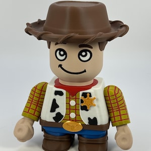May include: A toy figure of a cowboy with a brown hat, a red neckerchief, and a yellow sheriff's badge. The figure has a white vest with black cow spots, a yellow plaid shirt, and blue jeans. The figure is a collectible toy.