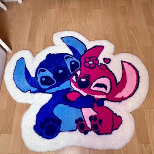 Pu&ograve; includere: Un tappeto in peluche con i personaggi Stitch e Angel del film d'animazione Lilo & Stitch. Stitch &egrave; blu, Angel &egrave; rosa e si abbracciano. Il tappeto ha un bordo bianco ed &egrave; posto su un pavimento in legno.