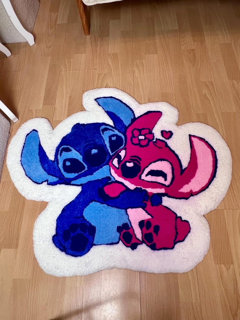 Pu&ograve; includere: Un tappeto in peluche con i personaggi Stitch e Angel del film d'animazione Lilo & Stitch. Stitch &egrave; blu, Angel &egrave; rosa e si abbracciano. Il tappeto ha un bordo bianco ed &egrave; posto su un pavimento in legno.