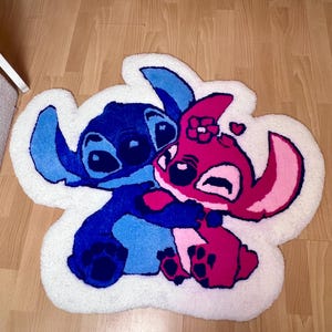 Pu&ograve; includere: Un tappeto fatto a mano con i personaggi Stitch e Angel di Lilo & Stitch. Il tappeto ha un bordo bianco e raffigura i personaggi in tonalit&agrave; di blu, rosa e magenta. Il tappeto &egrave; posto su un pavimento in legno.