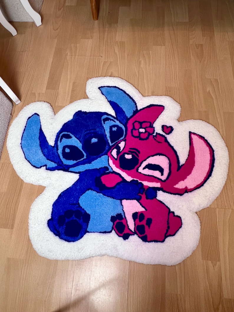 Pu&ograve; includere: Un tappeto fatto a mano con i personaggi Stitch e Angel di Lilo & Stitch. Il tappeto ha un bordo bianco e raffigura i personaggi in tonalit&agrave; di blu, rosa e magenta. Il tappeto &egrave; posto su un pavimento in legno.