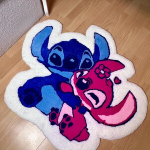 Pu&ograve; includere: Un tappeto bianco e morbido con l'immagine dei personaggi dei cartoni animati Stitch e Angel di Lilo & Stitch. Stitch &egrave; blu e Angel &egrave; rosa, entrambi con dettagli neri. Il tappeto ha un bordo bianco ed &egrave; posto su un pavimento in legno.