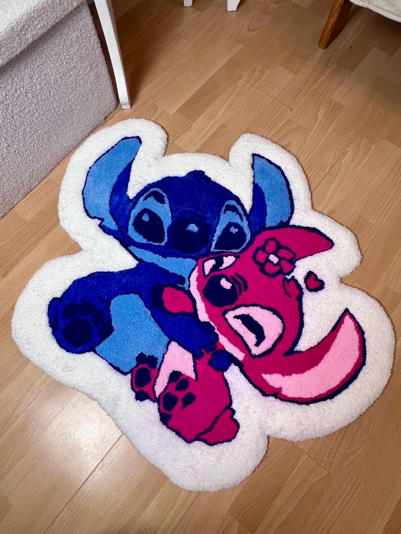 Pu&ograve; includere: Un tappeto bianco e morbido con l'immagine dei personaggi dei cartoni animati Stitch e Angel di Lilo & Stitch. Stitch &egrave; blu e Angel &egrave; rosa, entrambi con dettagli neri. Il tappeto ha un bordo bianco ed &egrave; posto su un pavimento in legno.