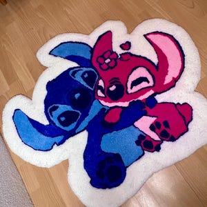 Pu&ograve; includere: Un tappeto fatto a mano con i personaggi Stitch e Angel di Lilo & Stitch. Il tappeto ha un bordo bianco e raffigura i personaggi in tonalit&agrave; di blu, rosa e magenta, che si abbracciano. Il tappeto &egrave; posto su un pavimento in legno.