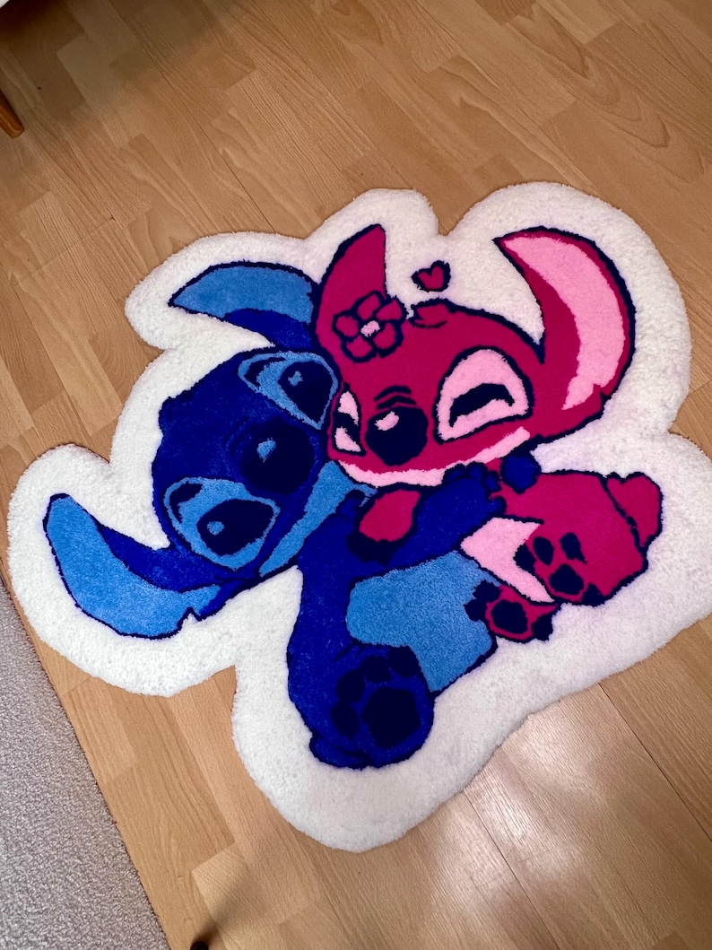 Pu&ograve; includere: Un tappeto fatto a mano con i personaggi Stitch e Angel di Lilo & Stitch. Il tappeto ha un bordo bianco e raffigura i personaggi in tonalit&agrave; di blu, rosa e magenta, che si abbracciano. Il tappeto &egrave; posto su un pavimento in legno.