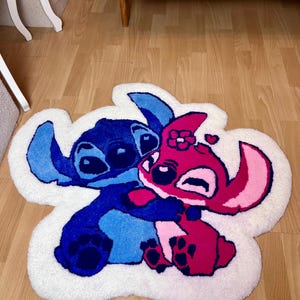 Pu&ograve; includere: Un tappeto bianco a forma di nuvola con un personaggio dei cartoni animati blu e rosa che si abbracciano. I personaggi si abbracciano, con il personaggio blu a sinistra e il personaggio rosa a destra. Il tappeto &egrave; su un pavimento di legno.
