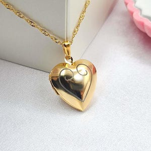 Puede incluir: Un collar de oro con un dije en forma de corazón y una cadena delicada. El dije tiene un diseño detallado y es perfecto para guardar una pequeña foto o mensaje.