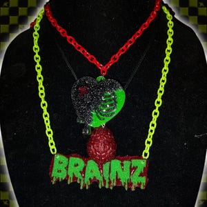 May include: A layered necklace set featuring a red plastic chain and a neon green plastic chain. The pendants include a glittery black and green heart with a ribcage design and a red glittery brain above a green dripping text plaque reading BRAINZ.