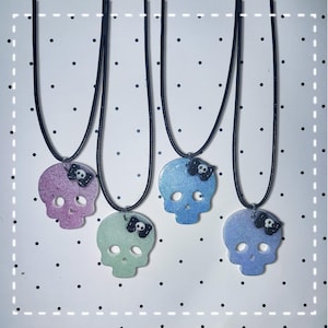 May include: Four skull-shaped pendants on black cord necklaces. Each pendant features a glittery finish in pastel purple, light green, light blue, or periwinkle. A small black bow with a tiny white skull center is attached to the top of each skull.