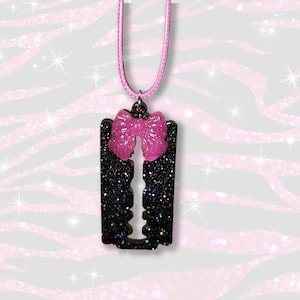 May include: A pendant necklace featuring a black glittery razor blade charm accented with a bright pink glittery bow. The charm hangs from a matching pink cord, creating a contrast between the dark, sparkling blade and the vibrant pink bow.