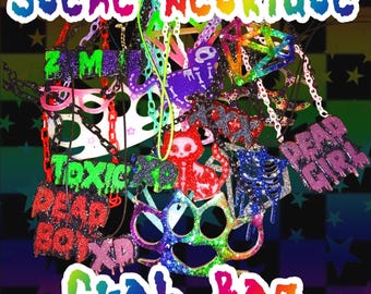 May include: A collection of colorful, glittery Scene Necklaces, including designs with text like "TOXIC" and "DEAD GIRL", and shapes such as a cat, brass knuckles, and a skeleton. The necklaces are on chains and cords, with the words "Grab Bag" at the bottom.