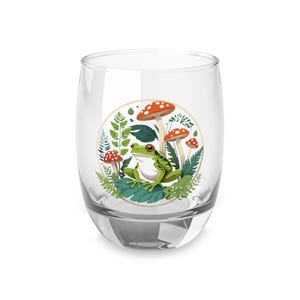 May include: A clear glass tumbler with a circular design featuring a green frog sitting on a bed of green leaves and red and white mushrooms.