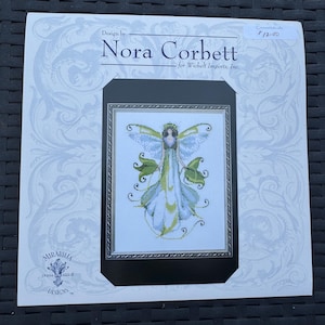 May include: A cross stitch pattern booklet titled Design by Nora Corbett for Wichelt Imports, Inc. The cover features a central illustration of a winged figure in a long, flowing blue and green gown, framed by a silver border on a white background.