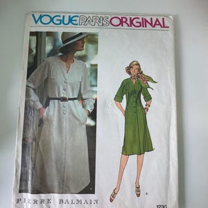May include: Vintage Vogue Paris Original sewing pattern featuring designs by Pierre Balmain. The pattern cover displays illustrations of two elegant dresses, one in a light color and the other in green. The text "Vogue Paris Original" is prominently displayed.