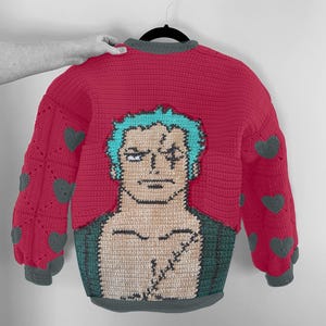 May include: A handmade red crochet sweater featuring a character design with teal hair and a scar. The sleeves have gray heart accents. The sweater has a gray collar and trim. A unique, cozy garment.