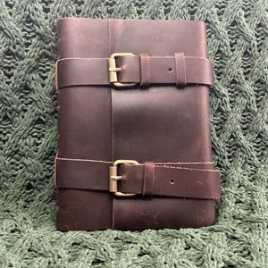 May include: A brown leather journal with two straps and brass buckles. The journal is closed and lying on a green knitted surface.
