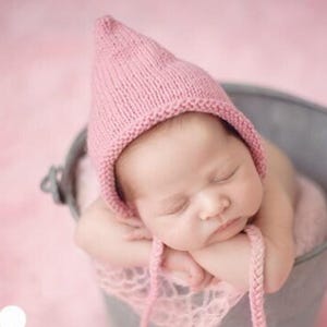 May include: A newborn baby wearing a pink knitted hat and wrapped in a pink blanket, resting peacefully inside a silver bucket. The background is a soft pink, creating a gentle and serene atmosphere. The image is from Little New York photography.