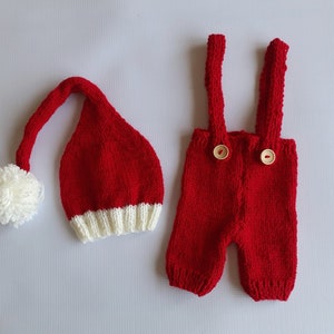 May include: A red knitted hat with a white pom-pom and a white ribbed brim, paired with red knitted overalls featuring two light-colored wooden buttons on the front straps. The set is crafted from a textured yarn in a vibrant solid red color.