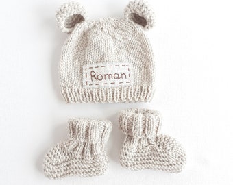 Könnte beinhalten: Eine beige gestrickte Baby-Mütze mit Bärenohren und einem Namensschild mit der Aufschrift "Roman". Die Mütze wird mit passenden beige gestrickten Booties geliefert.