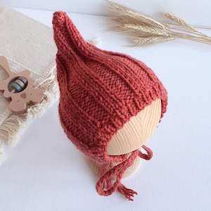 May include: A rust-colored, knitted baby bonnet with a pointed top and ribbed texture. The bonnet has a tie closure and is displayed on a wooden head form. The background includes a wooden toy and wheat stalks.