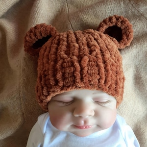 May include: A brown, textured knit baby hat with bear ears. The hat is worn by a baby wearing a white onesie. The baby has closed eyes and a peaceful expression. The background is a neutral beige.