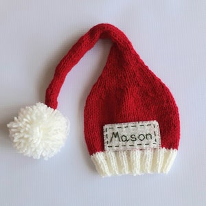 May include: Red and white knitted Santa hat with a white pom-pom and a name tag that says "Mason".