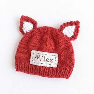 May include: Red knitted baby hat with white fox ears and a white patch that says "Miles" in brown embroidery.