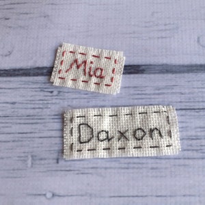 May include: Two white fabric labels with names embroidered on them. The top label says "Mia" in red thread. The bottom label says "Daxon" in black thread.