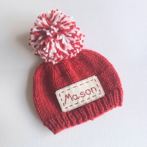 May include: Red knitted baby hat with a white pom pom and a white patch with the name "Mason" embroidered in red.