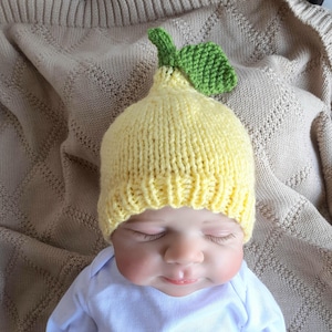 Puede incluir: Un gorro tejido a mano con forma de limón en color amarillo brillante, con un borde acanalado y una hoja verde texturizada unida en la parte superior. El gorro presenta un patrón de punto visible y un diseño de corona puntiaguda como un limón.