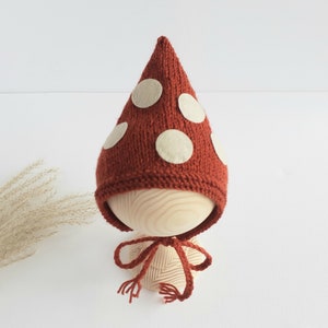 Può includere: Un cappello da neonato lavorato a maglia, a forma di fungo, color ruggine, con grandi pois in feltro color crema. Il cappello è legato con un cordino color ruggine abbinato. Il cappello è esposto su una forma di legno.