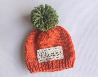May include: Orange knitted baby hat with a green pom-pom on top and a white patch with the name "Elias" embroidered on it.