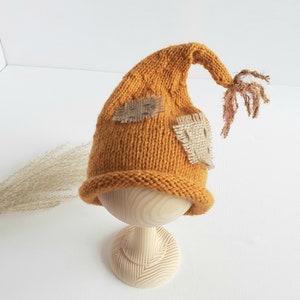 Op de afbeelding: Een mosterdgele gebreide muts met een puntige top en rafelige stoffen accenten. De muts wordt getoond op een houten standaard.