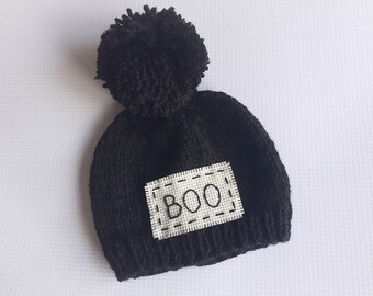 May include: Black knitted beanie with a fluffy pom pom on top and a white patch that says "BOO" in black embroidery.
